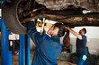 © CanBorg/peopleimages.com - Mechanic, woman and man under car in workshop for auto repair, service and inspection for safety. Engineering, partnership and teamwork for vehicle maintenance with tools, parts or industry at garage