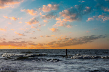 Beautiful and colorful silhouette of a surfer in the sun of dawn. Dramatic morning clouds over calm ocean waters with patient surfer. Puffy white clouds and warm morning sun