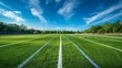 © Gia - A straight view down the middle of a football field with lush green grass and white marked lines, surrounded by trees and bathed in a clear blue sky with wispy clouds.