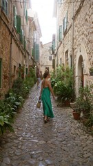 Poster - Woman walking in a narrow stone street of valldemossa, mallorca, spain, wearing a green dress on a sunny day with historic buildings and greenery around.