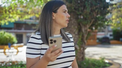 Wall Mural - Young attractive hispanic woman using smartphone standing in an urban park for a phone call, wearing a striped shirt, surrounded by city buildings and greenery.