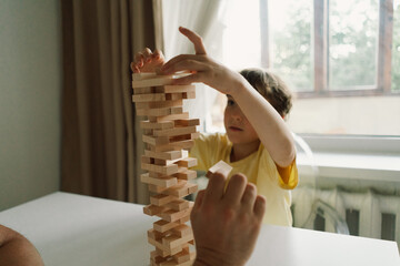 A father and son play a block tower game together indoors. The game is taking place on a white table, and the background is out of focus. Father's day