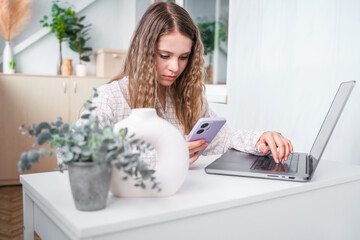 A young teenage student girl looking a laptop screen, online courses, studying remotely from home, sitting at a desk with a phone and laptop in a modern bright interior with plants.