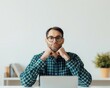 © Bantita - Man wearing a green plaid shirt sitting at a desk with a laptop, resting his chin on his hands in a contemplative pose.
