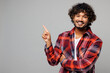 © ViDi Studio - Young happy smiling Indian man he wear red shirt casual clothes point index finger forefinger aside indicate on area mock up isolated on plain grey color background studio portrait. Lifestyle concept