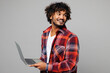 © ViDi Studio - Young happy smiling Indian IT man wear red shirt casual clothes hold use work on laptop pc computer look aside on area mockup isolated on plain grey color background studio portrait Lifestyle concept