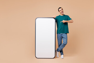 Canvas Print - Full body young middle eastern man wears blue t-shirt casual clothes point finger on big huge blank screen mobile cell phone smartphone with workspace area isolated on plain pastel beige background.