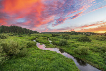 A winding river flows through a verdant meadow, bathed in the golden light of a beautiful sunset. The sky is ablaze with vibrant hues of orange, pink, and purple.