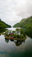 A small island with lush greenery and two red cabins sits in a calm, clear Norwegian lake surrounded by towering mountains. Lovrafjorden, Norway