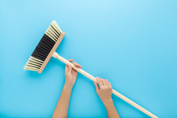 Young adult woman hands holding and showing new white black broom with wooden stick on light blue table background. Pastel color. Closeup. Cleaning tool. Point of view shot. Top down view.