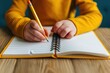 © Moopingz - A child wearing a yellow sweater writing in a spiral notebook with a yellow pencil on a wooden table.