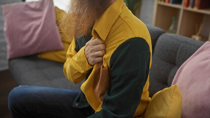 Wall Mural - A young bearded redhead man in colorful casual wear clutching his chest indoors on a couch with pillows.