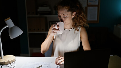 A young caucasian woman drinks coffee while working late in an office, illuminated by a desk lamp.