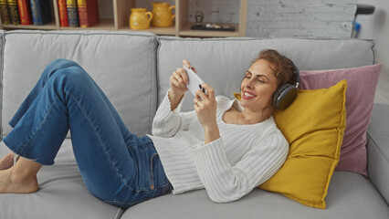 Wall Mural - Relaxed young woman in headphones gaming on smartphone on home couch, cozy casual lifestyle