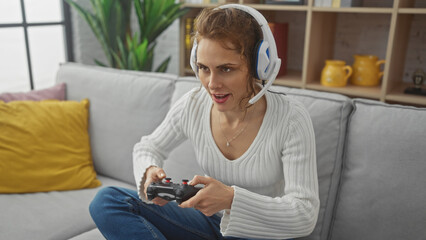 Wall Mural - A young woman engages in gaming sitting on a sofa at home, wearing headphones and holding a controller.