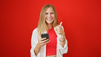 Sticker - Smiling blonde woman giving thumbs up while holding smartphone against red background.