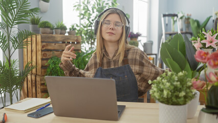 Sticker - A young, blonde woman wearing headphones enjoys music at her laptop in a cozy, plant-filled florist's shop.