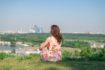 A young girl with a beautiful figure sits on the grass and looks at the city. The sky is blue and the grass is green.