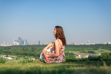 A young girl with a beautiful figure sits on the grass and looks at the city. The sky is blue and the grass is green.