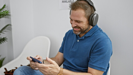 Wall Mural - Handsome middle-aged hispanic man with grey hair, smiling while using smartphone and headphones indoors.