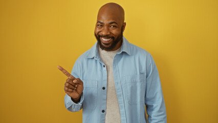 Handsome black man pointing against yellow background wall, wearing blue shirt and smiling boldly, embodying confidence and positivity in a visually striking portrait.