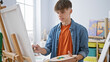 © Krakenimages.com - A young caucasian male artist focused on painting while holding a palette in a well-lit art studio