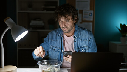 Poster - A young hispanic man in a denim jacket uses a smartphone while eating and working late in an office.