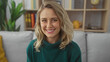© Krakenimages.com - A smiling young woman with blonde hair, dressed in a green sweater, sits comfortably in a cozy, bookshelf-decorated living room.