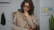 © Krakenimages.com - Young hispanic woman in an office, reviewing documents with concentration, wearing glasses and a ring, standing next to a colorful notice board and some office supplies.