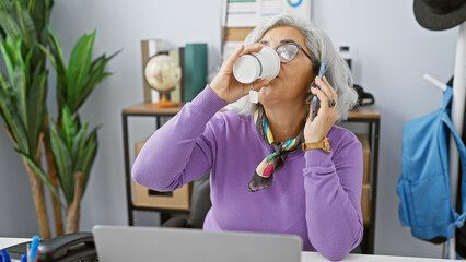 Poster - A mature woman multitasks in an office, drinking coffee and talking on the phone while seated at her desk.