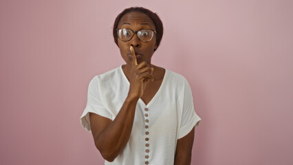 Wall Mural - Young african american woman making a shushing gesture over an isolated pink background wearing glasses and a white shirt