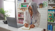 © Krakenimages.com - Elderly pharmacist woman with grey hair taking notes in a notebook while working in a pharmacy shop with a laptop