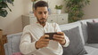 © Krakenimages.com - Handsome young hispanic man sitting indoors in a cozy living room at home, attentively looking at his phone while relaxing on a comfortable sofa.