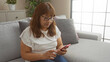 © Krakenimages.com - An elderly hispanic woman is seated on a couch in a bright living room, using a red smartphone.