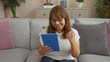 © Krakenimages.com - A middle-aged hispanic woman sitting on a sofa in her living room holds a tablet, smiling and giving a thumbs-up, capturing a moment of joy and connection in her cozy home.