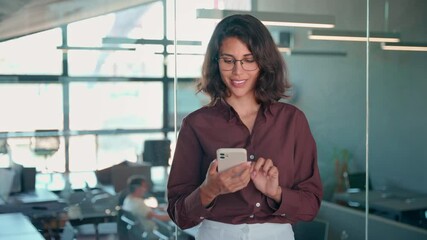 Wall Mural - Young dreaming Indian woman manager using cell phone mobile AI app for work call, business communication, trading. Pondering Latin Hispanic female businesswoman holding smartphone standing in office