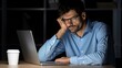 © lililia - A man wearing glasses sits at a desk lit by a desk lamp. He is working on his laptop with his hands on the keyboard