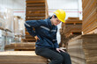 © Supachai - Female warehouse worker in a yellow helmet and blue uniform experiences back pain while sitting on stacked cardboard sheets at warehouse factory.