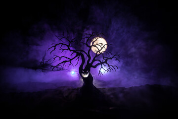 Silhouette of scary Halloween tree with horror face on dark foggy toned background with moon on back side. Scary horror tree with zombie and monster demon faces.