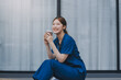 © ARMMY PICCA - A cheerful woman in scrubs holds a clipboard, standing confidently before a modern hospital building, representing professional healthcare service.