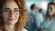 © svastix - A smiling woman with red hair and glasses stands out in an office environment, radiating positivity and professionalism while her colleagues work in the background, highlighting modern workplace cult