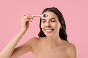 Wall Mural - Smiling woman applying makeup with brush on pink background