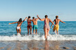 © Jose Calsina - Rear view of a group of young people having fun on the beach, running to the sea to take a dip in the water. Multiracial friends enjoying the summer vacation on the coast, taking a splash at the shore
