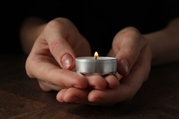 Wall Mural - Woman holding burning tealight candle at wooden table, closeup