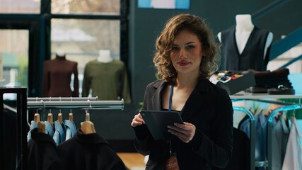 Wall Mural - Woman employee examining racks of clothes for stock inventory, using tablet to check all products. Retail store assistant checking discount list on device, ensuring commercial tags. Camera A.