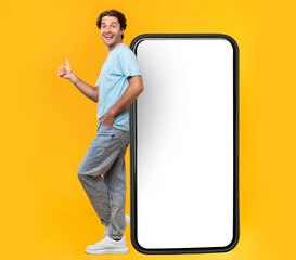 Wall Mural - Excited Man Leaning On Big Smartphone With Blank White Screen And Gesturing Thumb Up Sign, Cheerful Guy Recommending New App Or Website, Standing On Yellow Background, Mock Up Image, Full Body Length