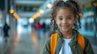 © Juanma Cuevas - 6 year old African American girl with orange backpack smiles in a school hallway. She is wearing a white t-shirt and a green jacket, representing back to school
