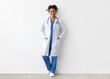 © Prostock-studio - Friendly female black doctor posing with hands in pockets looking at camera standing leaning on wall, lady wearing blue uniform, coat and stethoscope, full body length, white studio background banner