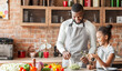 © Prostock-studio - Black smiling girl adding olive oil to veggies salad, dad mixing vegetables in bowl, kitchen interior, copy space
