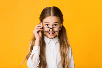 Wall Mural - Portrait Of Cute Schoolgirl Looking At Camera Over Eyeglasses On Yellow Studio Background.